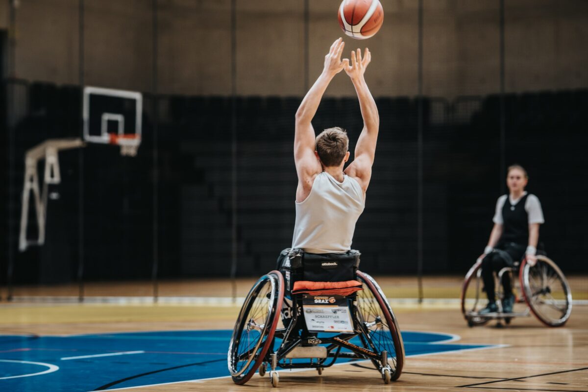 Ein Rollstuhlbasketballspieler in einem weißen Trikot fängt oder wirft einen Basketball in einer Sporthalle.