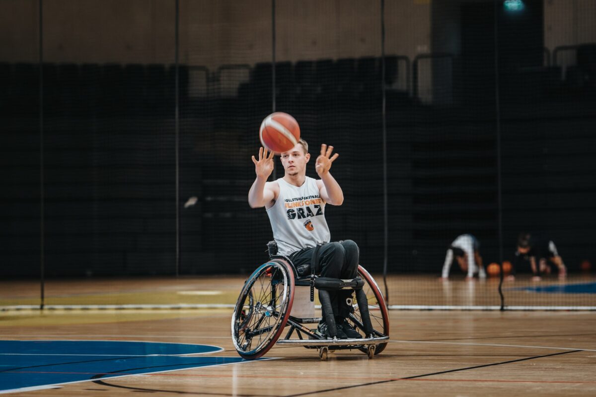 Ein Rollstuhlbasketballspieler in einem weißen Trikot mit der Aufschrift „GRAZ“ fängt oder wirft einen Basketball in einer Sporthalle.