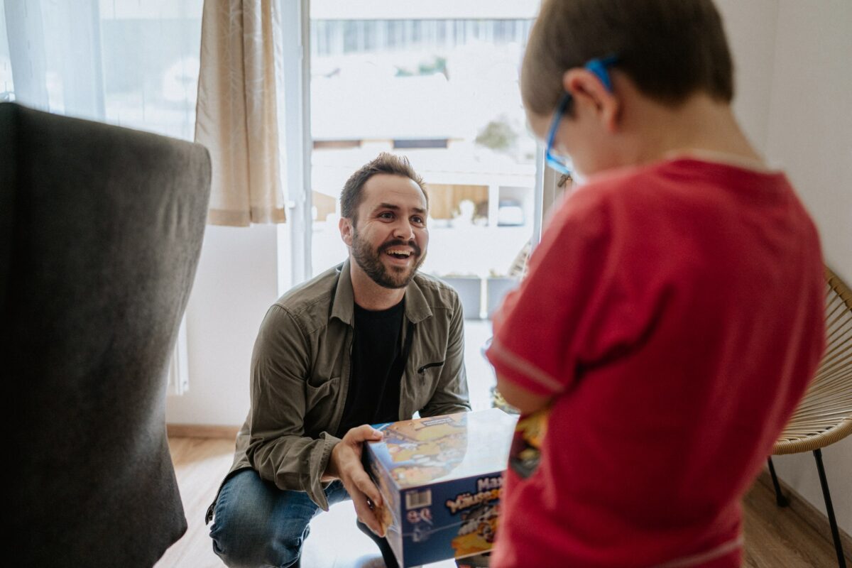 Marcel Resch sitzt lächelnd auf dem Boden und hält ein Spiel in der Hand, während er sich mit einem Kind in einem roten Shirt und blauer Brille unterhält.