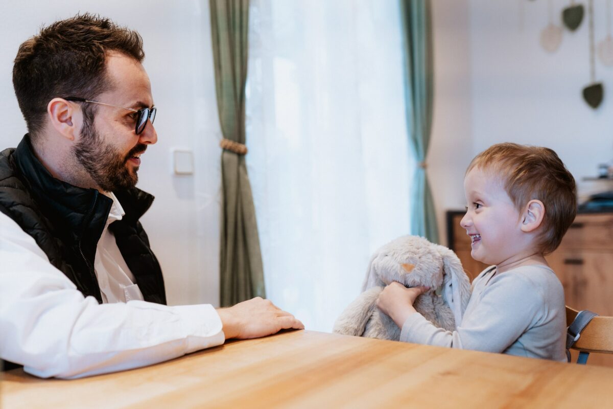 Marcel Resch sitzt lächelnd an einem Tisch und unterhält sich mit einem Kind, das einen grauen Stoffhasen umarmt.