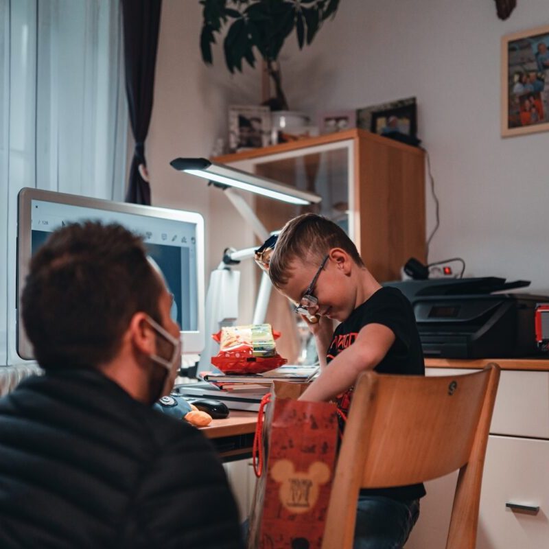 Niklas hält ein selbstgebautes Lego-Technik-Modell in der Hand und zeigt es Marcel Resch, der mit Maske im Hintergrund sitzt. Das Bild zeigt Niklas’ Weg zu mehr Selbstständigkeit durch Herz und Technik.
