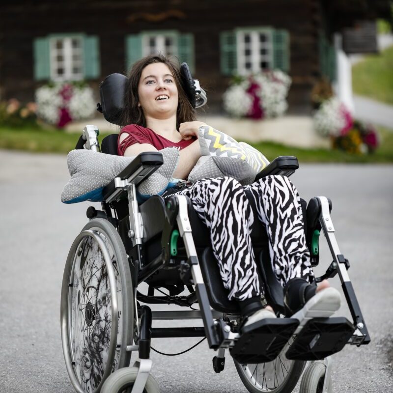 Eine lächelnde Frau im Rollstuhl mit gemusterter Decke auf den Beinen sitzt in einer natürlichen Umgebung mit Bergpanorama – Ein Unfall und alles ist anders: Laura’s Geschichte.