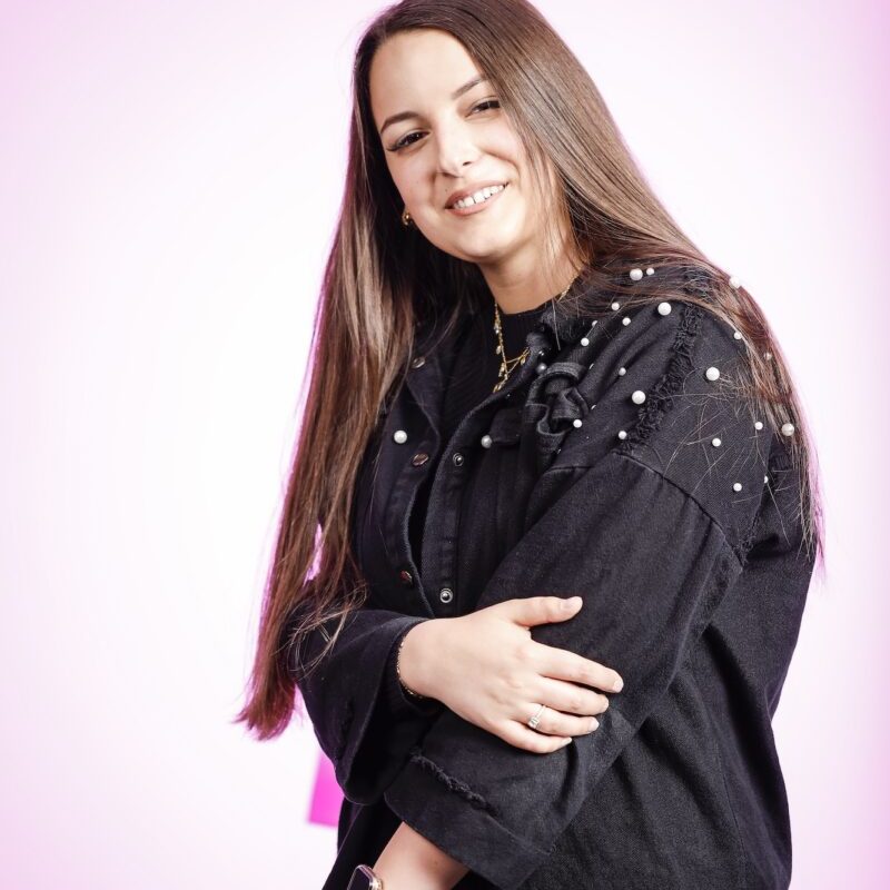 Porträtfoto von Angelina mit langen Haaren und schwarzem Oberteil auf pinkem Hintergrund für den Artikel ‚Shopping Tag für Angelina – Wenn der Krebs mal Pause macht‘