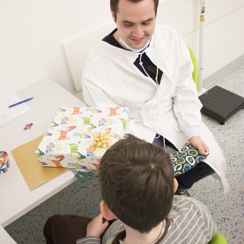 Marcel Resch in weißem Kittel und Maske überreicht einem Jungen im gestreiften Shirt ein bunt verpacktes Geschenk. Szene aus der Bilderserie „Von Lego, Hoffnung und Abschied: Eine besondere Freundschaft“.