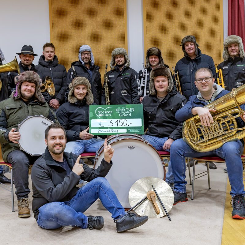 Eine Gruppe von Musikern mit ihren Instrumenten (Blasinstrumente und Schlagwerk) hält einen Spendenscheck über 3.150 € in die Kamera.