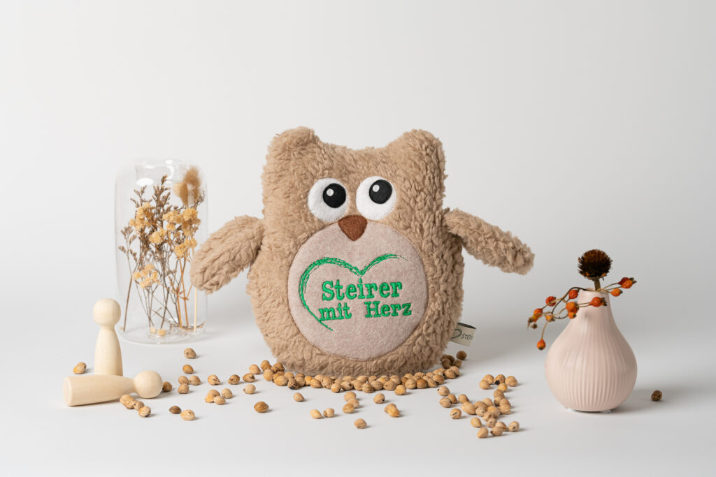 Ein Kirschkernkissen in Form einer kuscheligen Eule, auf dessen Ohr das grüne Logo „Steirer mit Herz“ aufgestickt ist. Es steht auf einem weißen Untergrund, umgeben von verstreuten Kirschkernen, kleinen Holzfiguren und Trockenblumen.