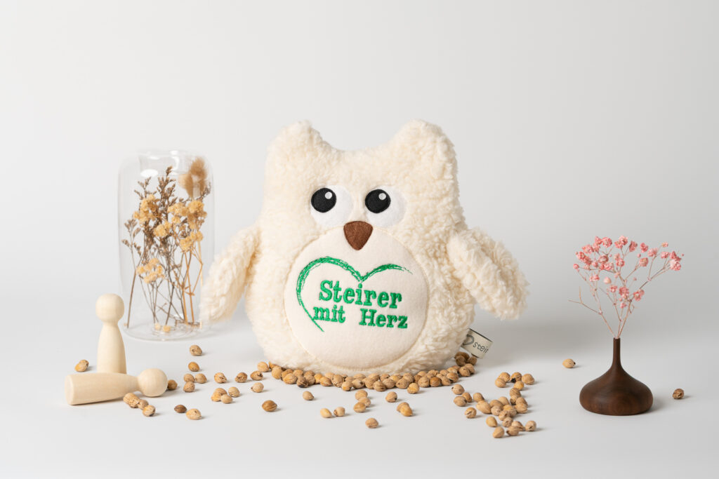 Ein Kirschkernkissen in Form einer kuscheligen Eule, auf dessen Ohr das grüne Logo „Steirer mit Herz“ aufgestickt ist. Es steht auf einem weißen Untergrund, umgeben von verstreuten Kirschkernen, kleinen Holzfiguren und Trockenblumen.