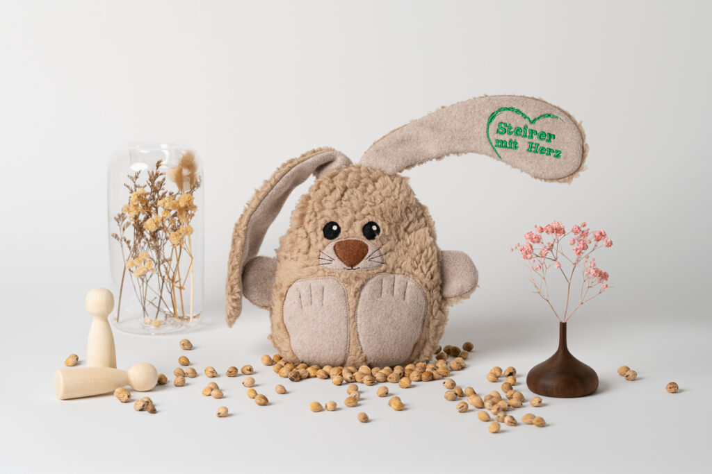 Ein Kirschkernkissen in Form eines kuscheligen Hasen, auf dessen Ohr das grüne Logo „Steirer mit Herz“ aufgestickt ist. Es steht auf einem weißen Untergrund, umgeben von verstreuten Kirschkernen, kleinen Holzfiguren und Trockenblumen.