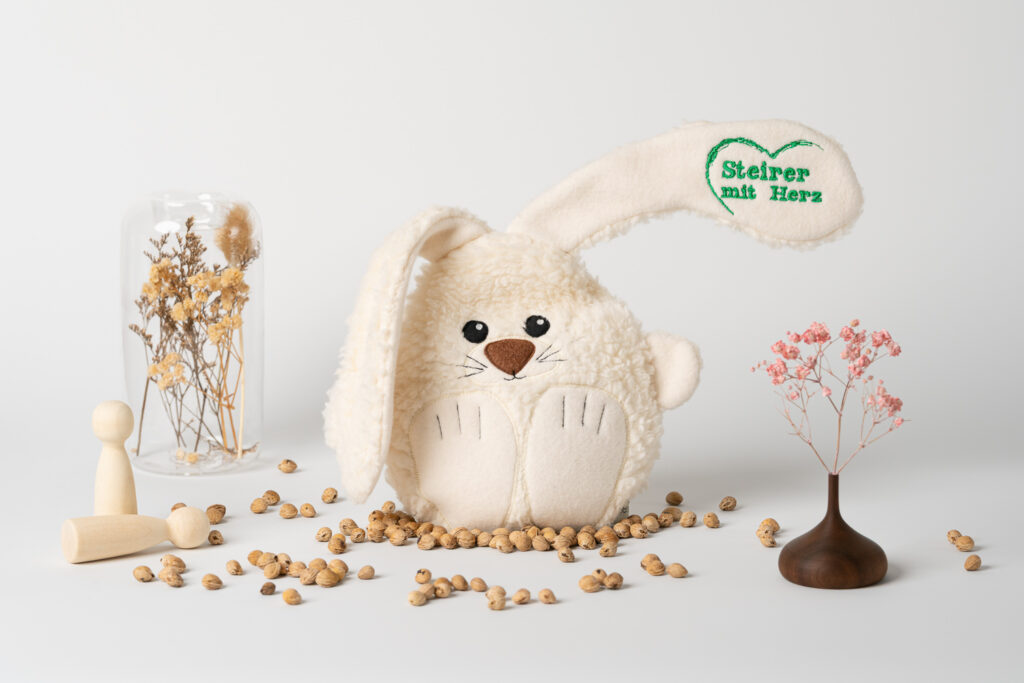 Ein Kirschkernkissen in Form eines kuscheligen Hasen, auf dessen Ohr das grüne Logo „Steirer mit Herz“ aufgestickt ist. Es steht auf einem weißen Untergrund, umgeben von verstreuten Kirschkernen, kleinen Holzfiguren und Trockenblumen.