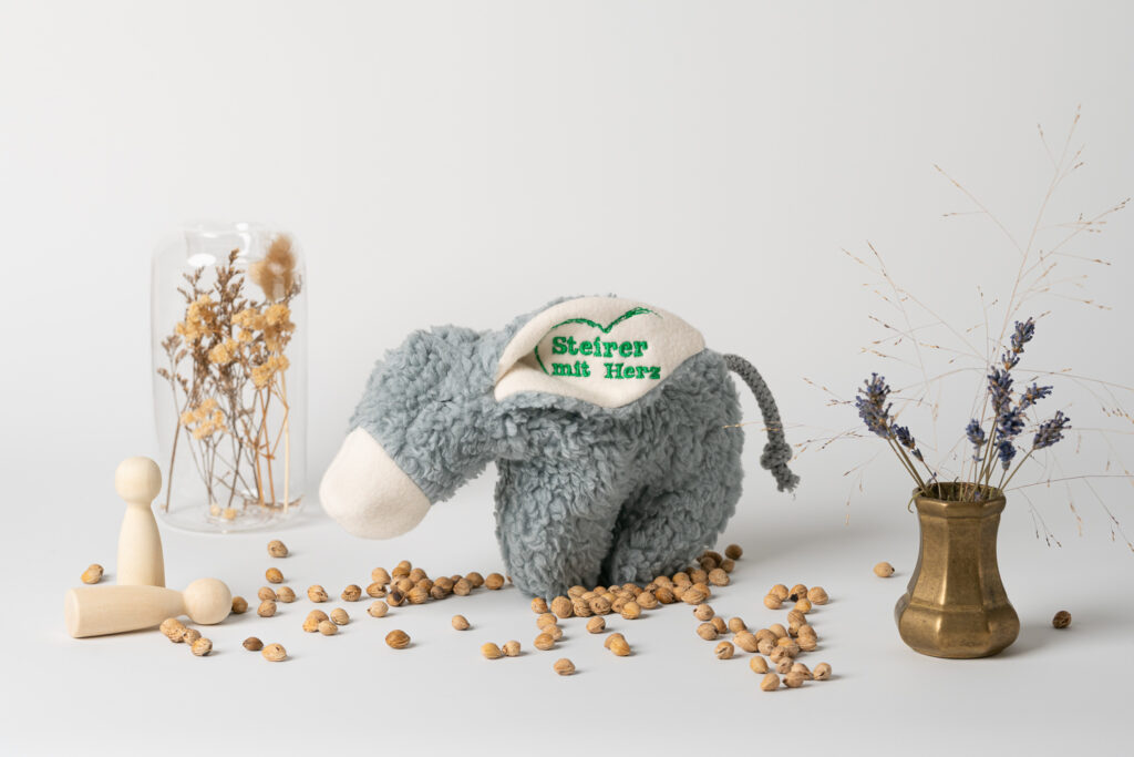 Ein Kirschkernkissen in Form eines kuscheligen Esels, auf dessen Ohr das grüne Logo „Steirer mit Herz“ aufgestickt ist. Es steht auf einem weißen Untergrund, umgeben von verstreuten Kirschkernen, kleinen Holzfiguren und Trockenblumen.