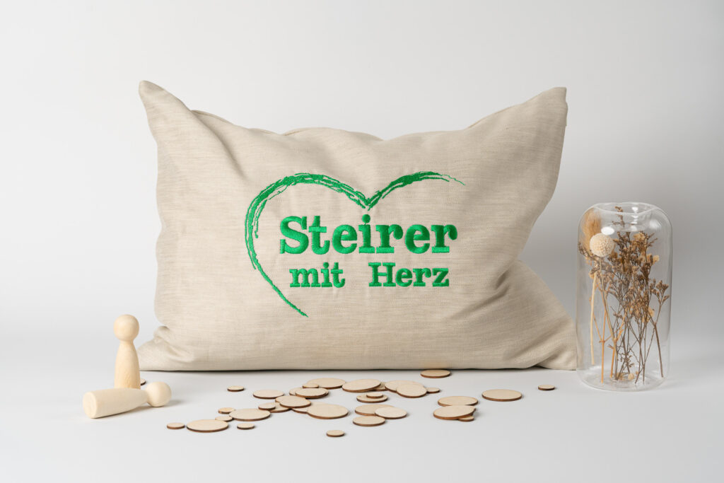 Ein beige-graues Kissen, auf dem in grüner Schrift das Logo „Steirer mit Herz“ aufgestickt ist, umgeben von kleinen Holzfiguren und einem Glas mit Trockenblumen.