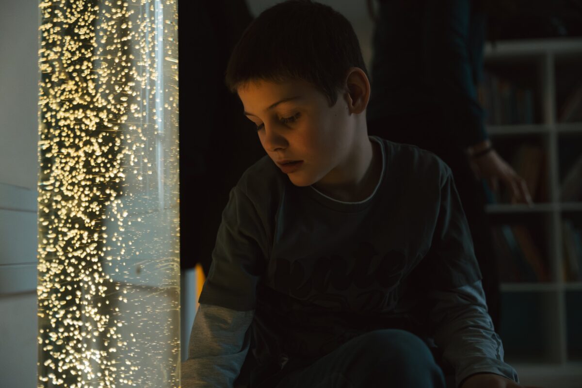 Ein Kind sitzt nachdenklich in einem Raum mit Lichtervorhang und blickt auf seine Hände. Das Bild zeigt eine Szene in der Volksschule St. Johann